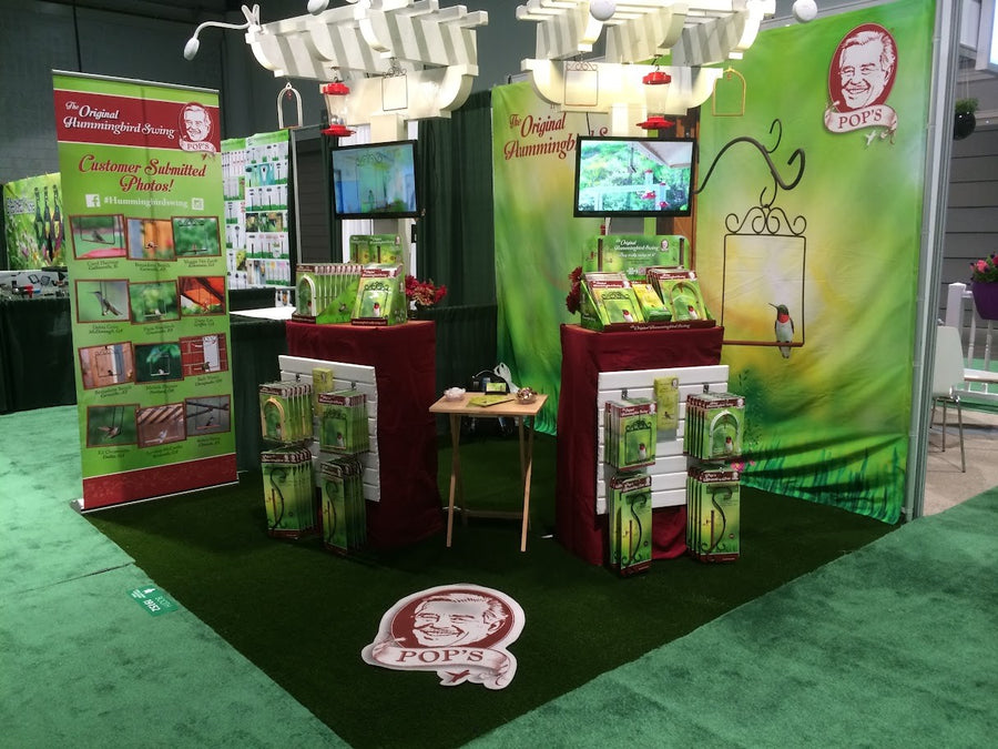 Pop's trade show booth in the green-package years, with a green hummingbird backdrop, white pergolas, swings on display and the Pops' face logo on the floor